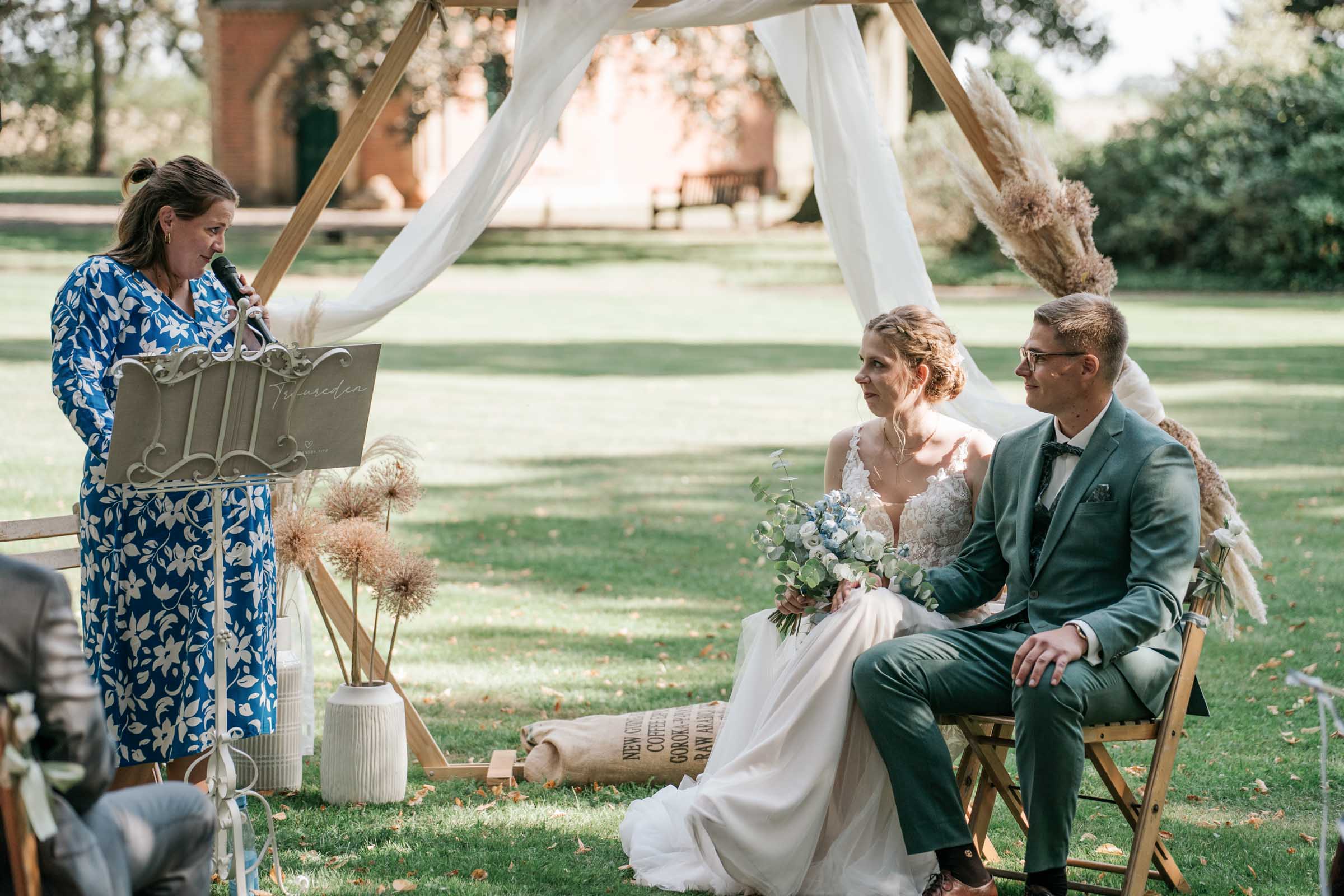 Traurednerin spricht, Brautpaar sitzt unter dem Traubogen im Grünen.