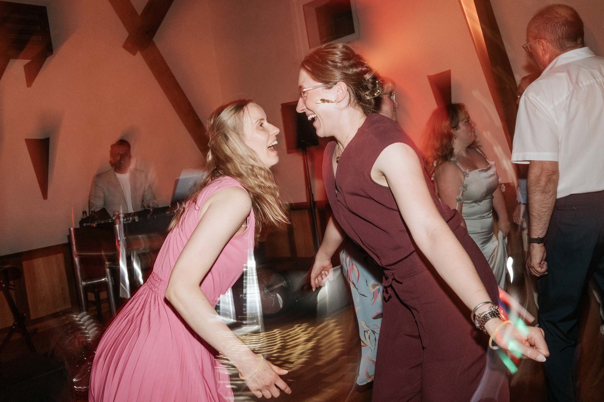 Zwei Frauen tanzen lachend; DJ im Hintergrund.