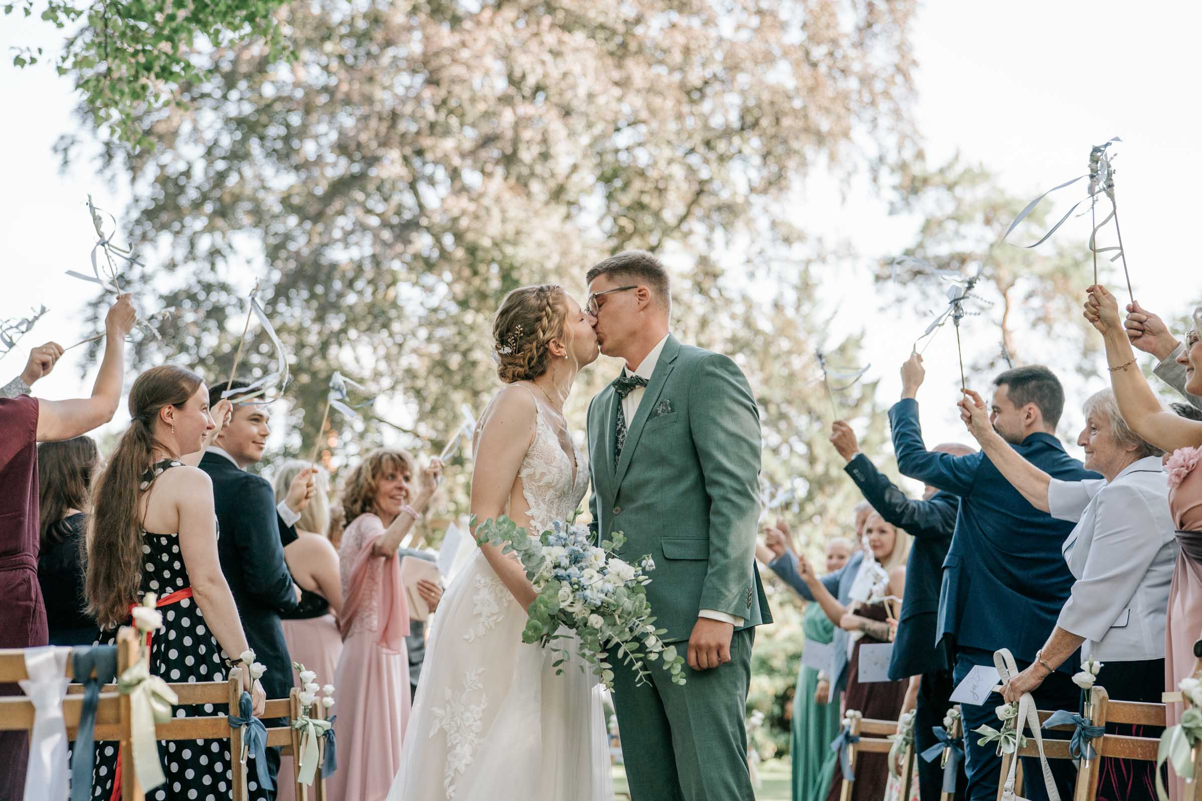 Brautpaar küsst sich auf dem Auszug, Gäste schwenken Bänder.